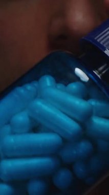 Dikey video. Beyaz ve Mavi Kapsüllerle Dolu Açık Mavi Şişe Bir Adamın Önünde, Hapların Dokusunu ve Parlak Sağlık Özelliklerini Vurguluyor