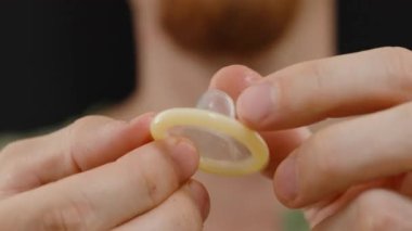 Arka planda bir erkek figürü ile iki el arasında tutulan bir prezervatifin yakın çekimi, uygun kullanım gösteren jest, lateks yüzeyine odaklanma ve şekil