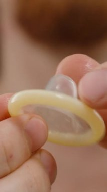 Dikey video. Arka planda bir erkek figürü ile iki el arasında tutulan bir prezervatifin yakın çekimi, uygun kullanım gösteren jest, lateks yüzeyine odaklanma ve şekil
