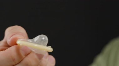 Arka planda bulanık bir erkek figürüyle prezervatif geren ellerin yakın çekimi, Hazırlık ve uygun tutuşu gösteriyor