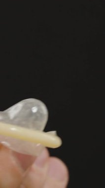 Dikey video. Arka planda bulanık bir erkek figürüyle prezervatif geren ellerin yakın çekimi, Hazırlık ve uygun tutuşu gösteriyor