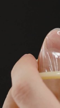Dikey video. Siyah bir zemin karşısında parmağına prezervatif takan bir adam, Doğru Tekniği ve Kullanımı Ellere ve Kondoma Odaklı Gösteriyor