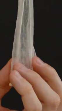 Dikey video. Siyah Arkaplan Karşısında Parmaklar Arasında Tutulan, Uzunluğunu ve Şeklini Vurgulayan Tamamıyla Açık Bir Kondom