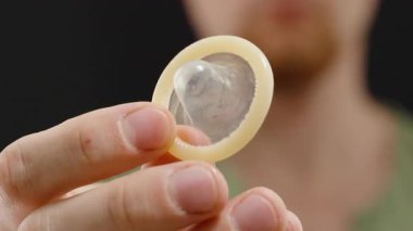 Siyah Arkaplan Karşısında Kameraya Yakın Kondom Tutan Bir Adam Tasarım ve Fonksiyonunu Vurguluyor