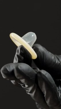 Dikey video. Siyah Eldivenli Bir El Parmaklar Arasında Bir Kondom Çimdikliyor, Uygun Tutuş ve Güvenliği Vurguluyor