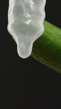 Dikey video. Yeşil bir salatalığın yakın çekimi kısmen şeffaf lateks prezervatifle kaplıdır. Siyah arka planda, çarpıcı ışıklandırma güvenli seks eğitimi içeren desen ve detay gösterilir.