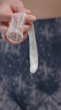 Dikey video. İçinde meni olan kullanılmış bir prezervatif tutan bir kadın. İçindekilere dokunuyor. Güvenli seks uygulamalarını ve doğum kontrolü kavramını vurguluyor.