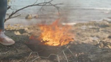 Rocky Shoreline 'da, Portatif Izgara' da Küçük Kamp Ateşi Parıldıyor. Arka planda Okyanus ve Gündelik Ayakkabılı Bir Kişinin Kısmi Görünümü.