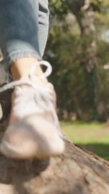 Dikey video. Pembe spor ayakkabı giyen bir kadın güneşli bir parktaki ağaç gövdesine dikkatle basıyor. Etrafı yeşil otlar, gölgeler ve doğal orman manzaralarıyla çevrili.