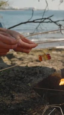 Dikey video. Arka planda okyanus manzaralı bir aile pikniğinde üç el marşmelov ve sakızları bir kamp ateşi üzerinde kızartıyordu.