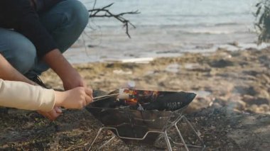 Üç kişi, muhtemelen çocuklu bir aile, deniz kenarındaki küçük bir şöminede şekerleme ve sakız kızartıyorlar, manzaralı rahatlatıcı bir pikniğin tadını çıkarıyorlar.