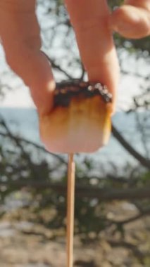 Dikey video. Yangında kavrulmuş bir marşmelovun kızarmış kabuğunu soyan bir kadın, arka planda ağaçlar ve okyanusla yumuşak erimiş merkezi açığa çıkarıyor.