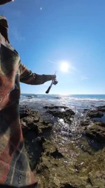 Parlak güneşli bir gökyüzünün altında dalgaların çarptığı kayalık bir deniz kıyısında elinde olta tutan ve arka planda parıldayan bir okyanus ufkunu gören ilk kişi.