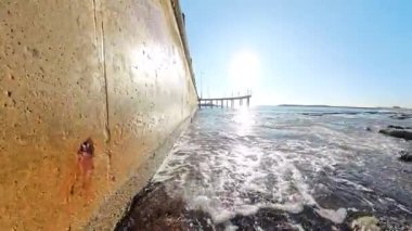 Beton bir duvarın yakınındaki sudan yansıyan düşük açılı güneş ışığı arka planda açık mavi bir gökyüzü ve kayalık bir kıyı şeridi olan bir rıhtıma çıkıyor.
