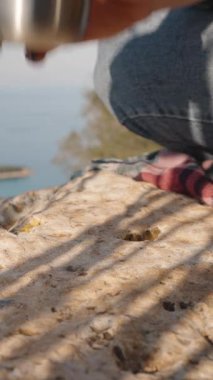 Dikey video. Güneş Işığı ve Gölgeli Kıyı Manzarasına Bakan Kayalık Yüzeyde Metal Bir Kupa Tutan Kadın Açık Hava Anı 'nı temsil ediyor.