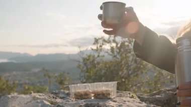Şapka takan ve güneş gözlüğü takan, Metal Kupa 'dan çay yudumlayan, Arkasında Güneş Parlayan,