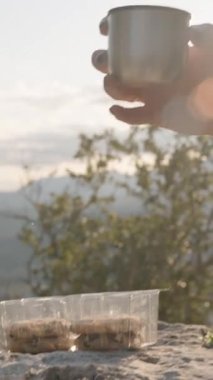 Dikey video. Şapka takan ve güneş gözlüğü takan, Metal Kupa 'dan çay yudumlayan, Arkasında Güneş Parlayan,