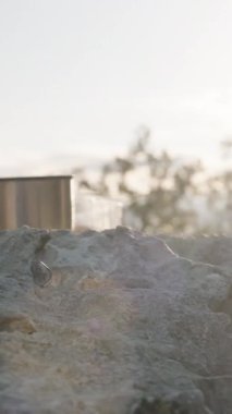 Dikey video. Sıcak Çayın Buharlı Kaya yüzeyinde Termos ve Manzaralı Dağ ve Deniz Manzaralı Sunset 'te Huzurlu Açık Hava Anı' nı sunar.