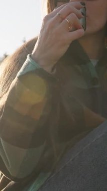 Dikey video. Bir Metal Kupa 'dan sıcak çay yudumlayan, doğa ve ağaçlarla çevrili, sakin bir açık hava anını temsil eden, kep ve güneş gözlüğü takan bir kadın.