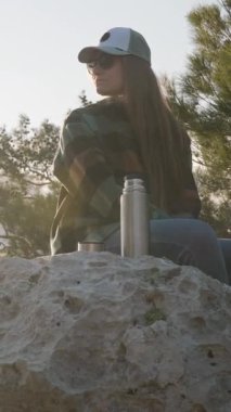 Dikey video. Bir termos ve bir fincan sıcak çayla büyük bir kayanın üzerinde oturan, etrafındaki ağaçlarla uzaklığa bakan, doğada yalnızlık anını temsil eden bir kadın.