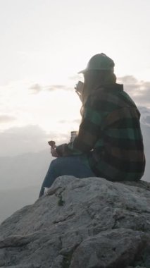 Dikey video. Siluetli Kadın Kayalık Zirvede Oturuyor Bulutlu Günbatımına Karşı Bir Çay Kadehi Tutuyor Doğada Bir Yalnızlık ve Yansıma Anını Temsil Ediyor