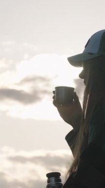 Dikey video. Termostan sıcak çayı bardağa döken elinde bir dağ günbatımına karşı bir kurabiye tutan kupadan yükselen buhar, sıcak bir açık hava anını temsil eden bir kadın.
