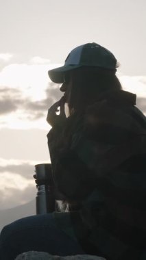 Dikey video. Bir Kaya 'da Oturan Sıcak Çay İçen Kadın Karlı Dağ Tepelerinde Gün Batımını İzlerken Kupadan Buhar Yükseliyor, Huzurlu Doğadan Kaçışı Temsil Ediyor