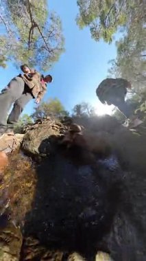 Dikey video. Temiz Su ve Güneş Işığı Yansımaları ile Rocky Nehri 'nden Geçen İki Gezgin Düşük Açı Görünümü