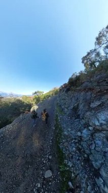 Dikey video. İki Gezgin Dik Dağ Yolu 'nda, Orman Tepeleri' nin Panoramik Manzarası ve Uzak Kıyı Kasabası 'yla yürüyor.