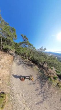 Dikey video. İki Yürüyüşçü, Yeşil Dağ Vadisi Manzarası ile çevrelenmiş Rocky Yolu boyunca yürüyor. Mavi bir gökyüzü altında, Güneş tepeden parlıyor.