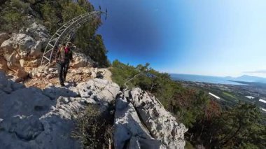Deniz, sahil ve şehir manzaralı, ağaçlar ve açık mavi gökyüzü ile çevrili Yüksek bir dağda Büyük Kayalıkların Üzerinde Tırmanan Adam