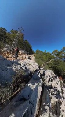 Dikey video. Deniz, sahil ve şehir manzaralı, ağaçlar ve açık mavi gökyüzü ile çevrili Yüksek bir dağda Büyük Kayalıkların Üzerinde Tırmanan Adam