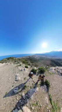 Dikey video. Bereketli yeşil bir vadiye bakan bir dağın tepesinden geniş bir hava manzarası ve açık mavi gökyüzü ile deniz ve yukarıda parlayan güneş.