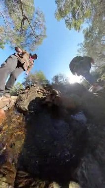 Dikey video. Temiz Su ve Güneş Işığı Yansımaları ile Rocky Nehri 'nden Geçen İki Gezgin Düşük Açı Görünümü