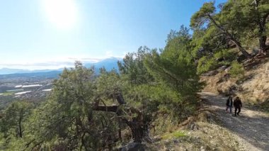 Aşağıdaki sahil ve şehir manzaralı yeşil ağaçlarla çevrili Rocky Dağı 'nın tepesinde yürüyüş yapan erkek ve kadın onları takip eden hareketli bir kameradan görüntülendi.