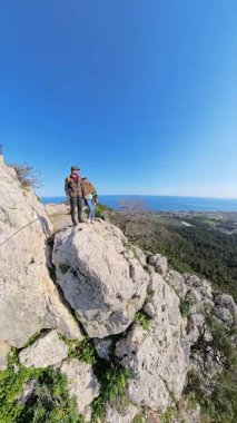 Dikey video. Kayalık bir uçurumun kenarında dikilen ve nefes kesen bir dağı ve güneşin altındaki deniz manzarasını seyreden çift el ele tutuşuyor.
