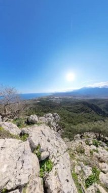 Dikey video. İki Yürüyüşçü Dağ Tepesi 'ndeki Büyük Kaya' nın üzerinde duruyor. Çevresi yeşillik ve berrak mavi gökyüzüyle çevrili Vadi ve sahil manzarası çarpıcı.