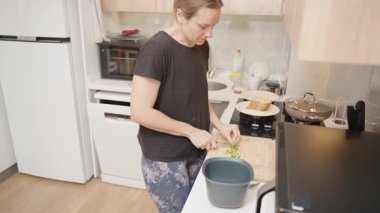 Siyah tişörtlü kadın doğranmış yeşil sebzeleri tahta kesme tahtasından modern mutfaktaki plastik konteynıra aktarıyor.