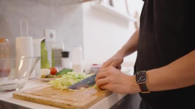 Mutfak tezgahında cam bir kaseye doğranmış Marul Düşüren Eller 'in yakın çekimi, Taze İçindekiler ve Yemek Utensilleri ile çevrili, Bir Salata Hazırlama