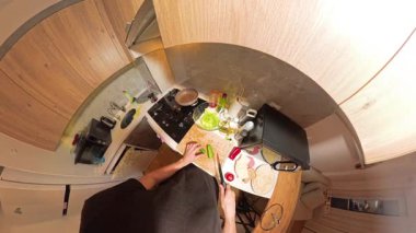 Küçük bir mutfaktaki ahşap kesim tahtasında salatalık kesen bir insanın 360 derecelik birinci derece görüntüsü, Taze İçindekiler ve Yemek Utensilleri ile çevrili.