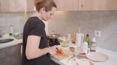 Domatesleri doğrama tahtasından cam bir kaseye aktaran, mutfak tezgahında mevsimler ve pişirme malzemeleriyle çevrili ellerin yakın çekimi.