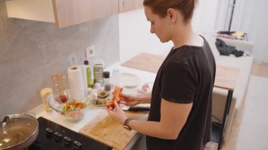 Modern bir mutfakta kırmızı biber tutan kadın ahşap kesim tahtasındaki taze salata için malzemeleri hazırlıyor çeşitli mutfak gereçleri ile çevreleniyor.