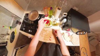 Tahta kesim tahtasında domates doğrayan bir insanın 360 derecelik birinci derece görüntüsü, Küçük bir mutfakta taze malzemeler ve mutfak gereçleri ile çevrili.