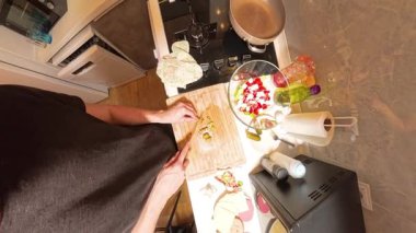 Tahta kesim tahtasında yeşil zeytin doğrayan bir kişinin 360 derecelik birinci derece görüntüsü, Küçük bir mutfakta çeşitli taze malzemelerle salata hazırlamak.