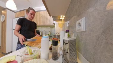 Modern bir mutfakta, tezgahta çeşitli yemek malzemeleriyle ahşap kesim tahtasının üzerinde peynir bloğu kesen kadın.