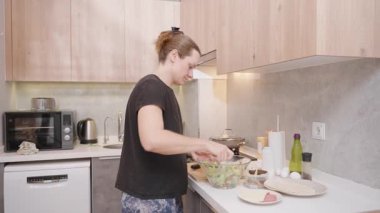 Modern Kitchen 'daki Cam Kasede Siyah Gömlekli Kadın Ahşap Dolaplar ve Tezgahta Çeşitli Malzemeler ile Karıştırma Salatası. Ev Yemekleri Konsepti
