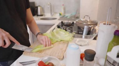 Modern bir mutfağın ahşap kesim tahtasındaki taze marul kesen kadın etrafını sebzeler, baharatlar ve yemek takımları sarmış sağlıklı bir salata hazırlıyor.