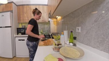 Siyah Gömlekli Kadın, Modern Kitchen 'da beyaz tabağa taze salata servis ediyor. Yanında tahta dolaplar ve tezgahta çeşitli malzemeler var. Sağlıklı Yeme Konsepti