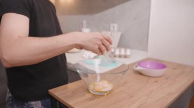 Bir cam kaseye yumurta sarısı, ahşap mutfak tezgahına yumurta sarısı, yemek tarifi kitabı ve arka plandaki yumurtalarla şeker döken kadının yakın çekimi.