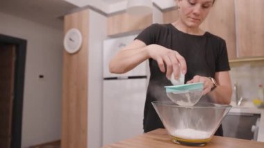 Tahta mutfak tezgahında yumurta sarısı ve şekerli cam bir kâseye un yerleştiren kadın, küçük bir elek kullanıyor.